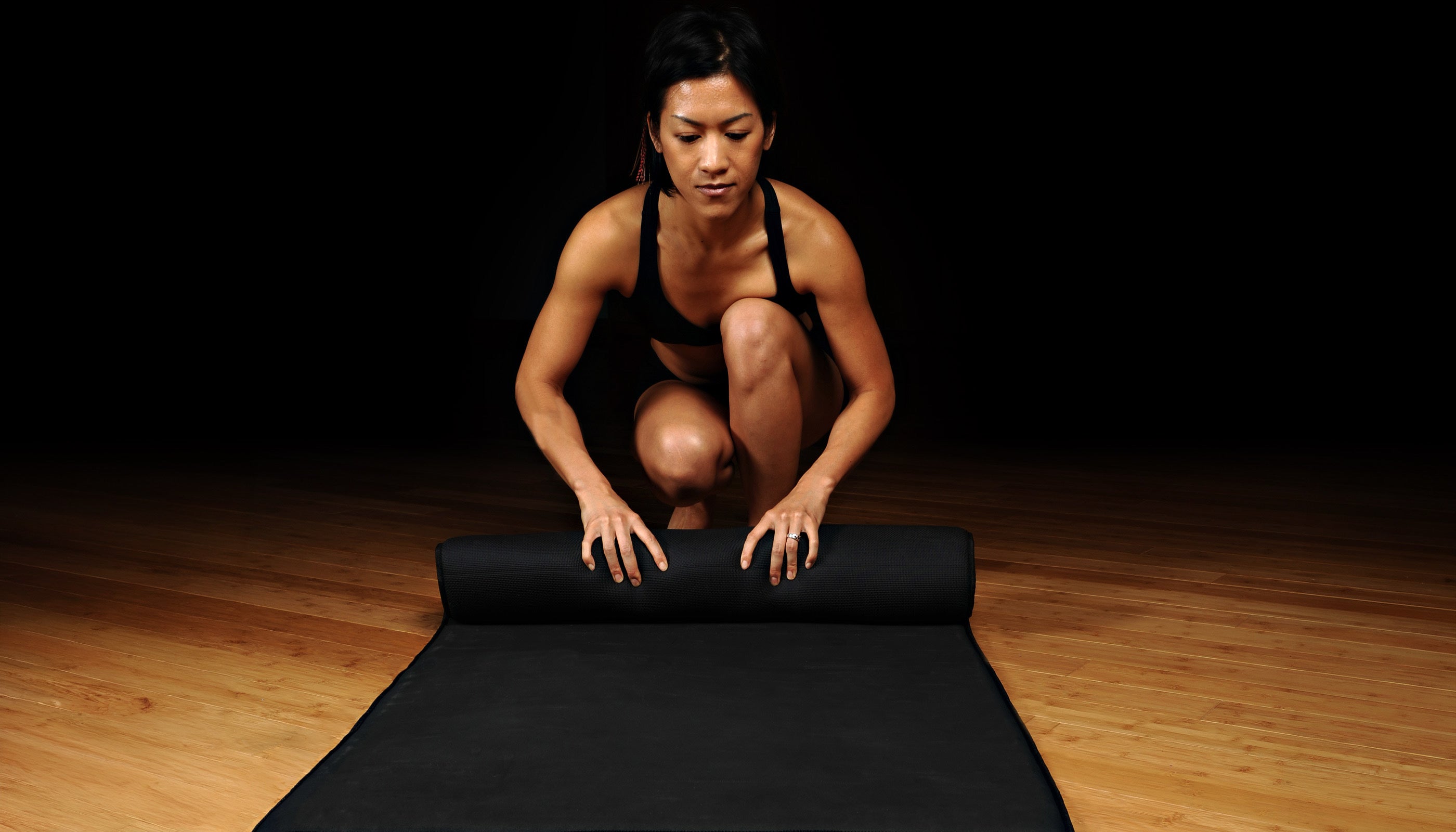 Woman crouching on a hardwood floor, rolling up a thick black yoga mat